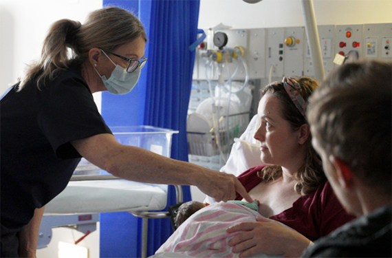 A midwife wearing a mask checks a baby being held by it's mother. The mother is sitting in a hospital bed and her partner is looking on.