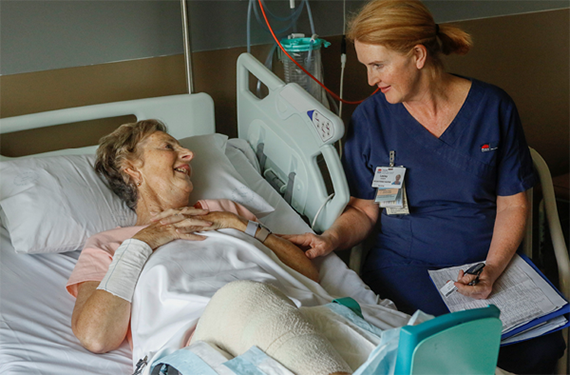 A mature-aged woman wearing a NSW Health registered nurse uniform, sitting next to an elderly woman in a hospital bed, actively listening and comforting her.