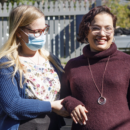 A woman with blond hair, wearing a mask, supporting a young woman with dark hair and glasses. by the arm