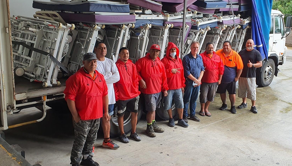 8 Tongan men standing in front of a truck full of hospital equipment, including beds, mattresses and trolleys.