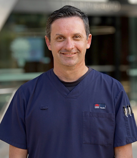 A man with short dark hair, smiling, wearing dark blue NSW Health scrubs.