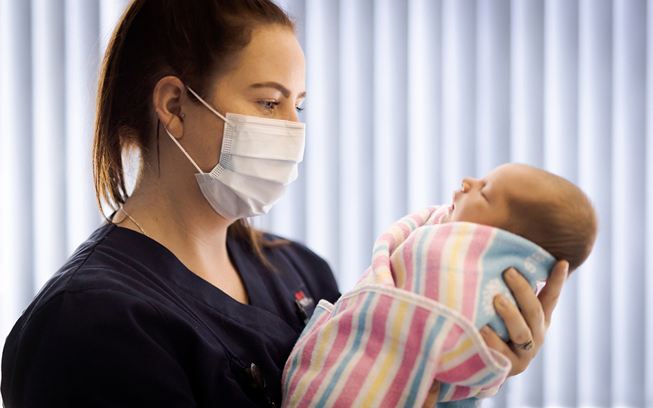 A midwife in a mask, holding a baby wrapped in a striped blanket.