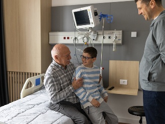 A child sitting on a family member’s lap. Beside them is another family member. They are in a hospital room.