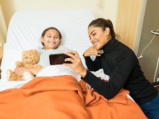 A child resting in a hospital bed while another person shows something on a phone.