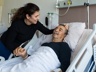 An older person resting in a hospital bed and family member holding their hand next to them.