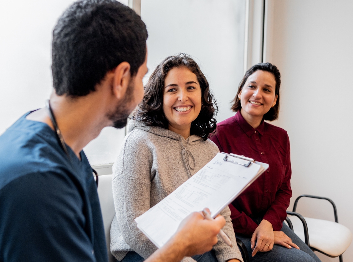 A guardian supporting someone else in a meeting with a doctor who is holding a clipboard.