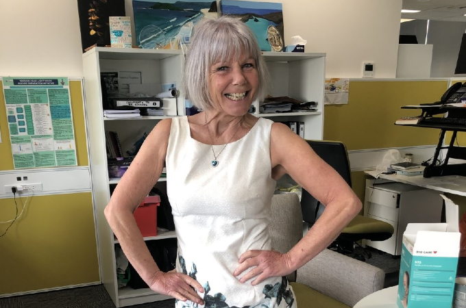 A woman with blonde-grey hair wearing a white dress has a big smile on her face and has her hands on her hips
