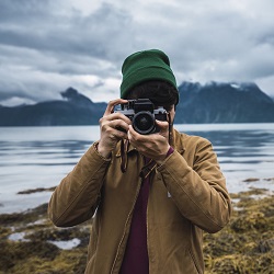 Man in beanie taking a photo next to a lake on an overcast day